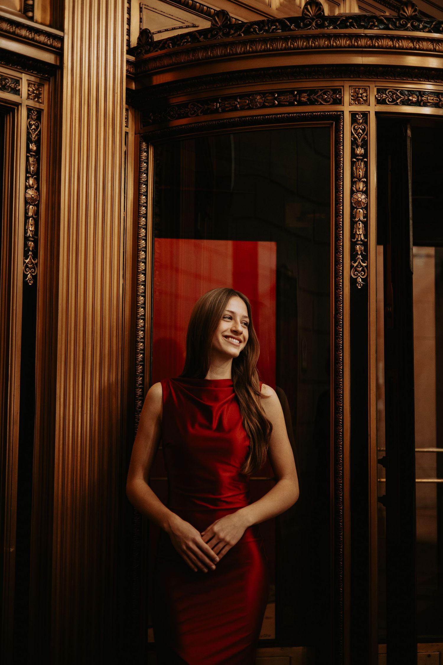 Woman in red dress poses elegantly against ornate gold-framed mirrors in dramatic lighting.