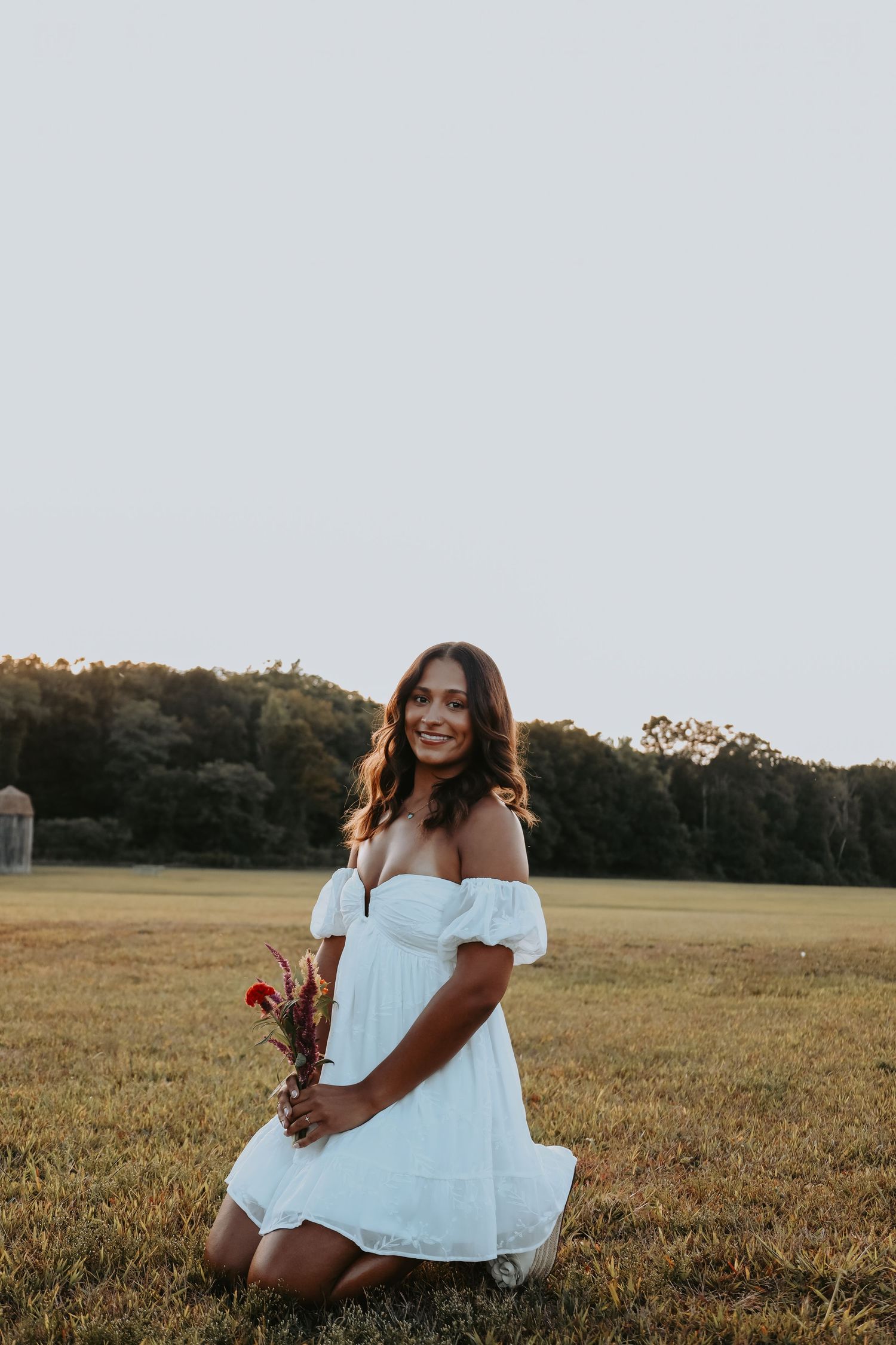 A person in a white dress sits in a grassy field during golden hour at sunset.