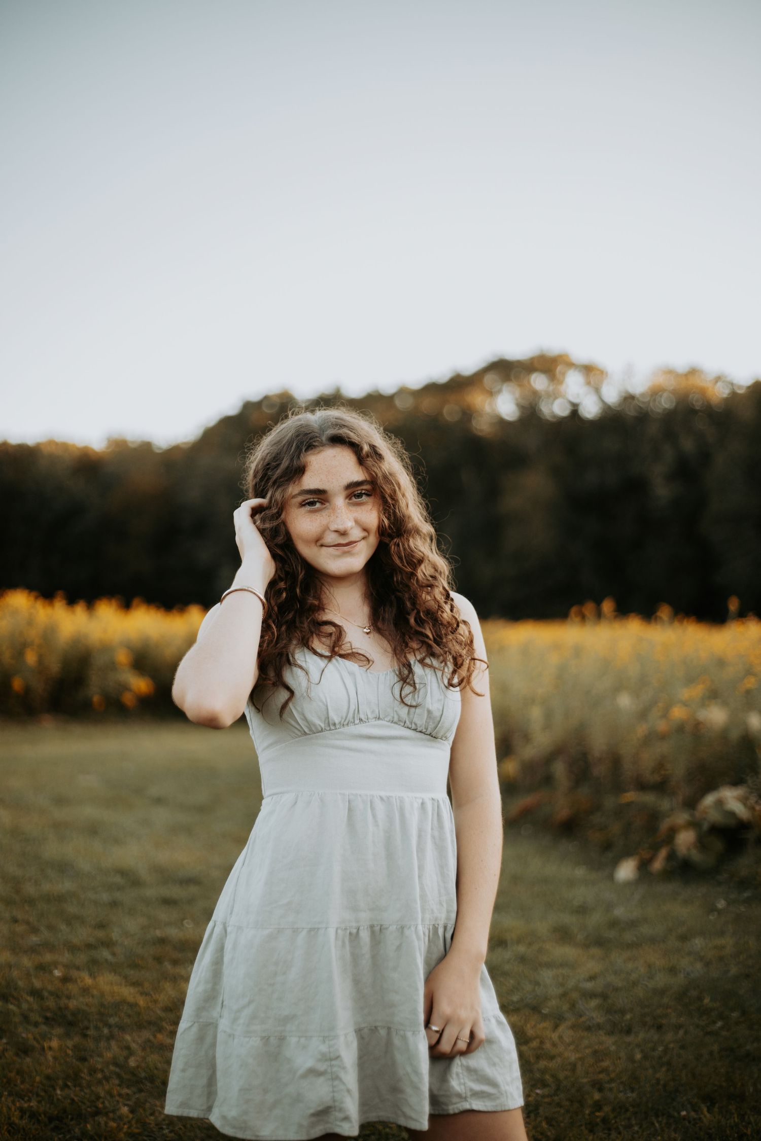 A person in a white flowing dress stands in a golden field at sunset with trees in the background.