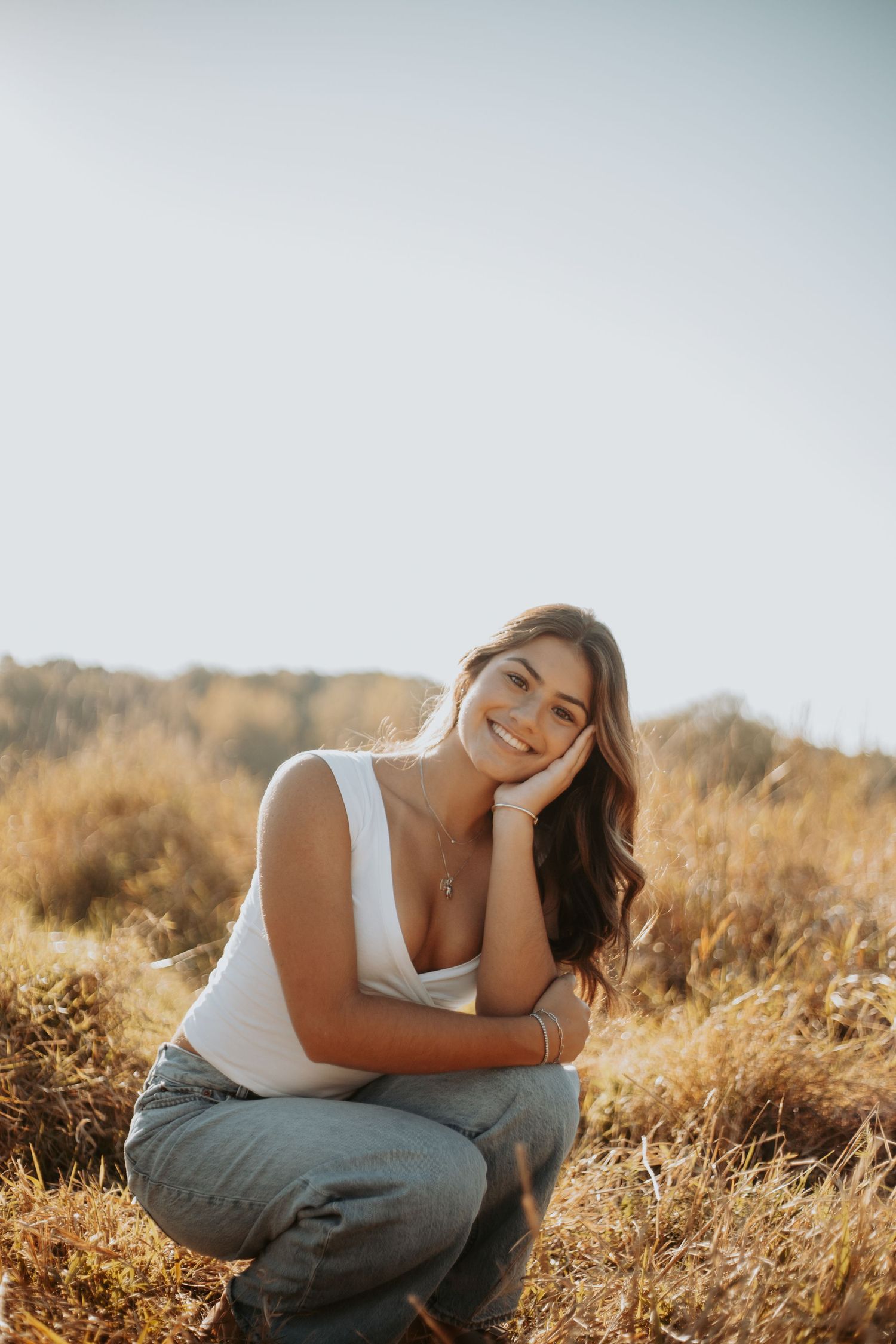 A person in a white tank top and jeans sits in a golden field at sunset with a warm glowing atmosphere.