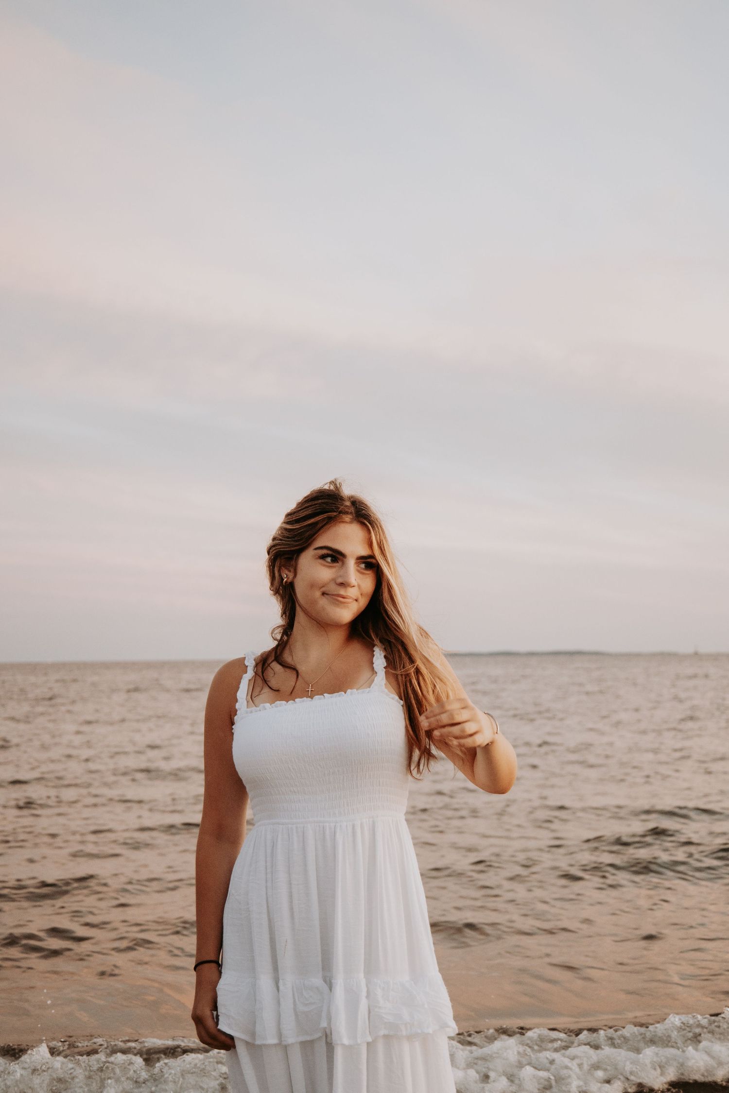 Woman in flowy white dress stands on beach at sunset with waves and ocean in background.