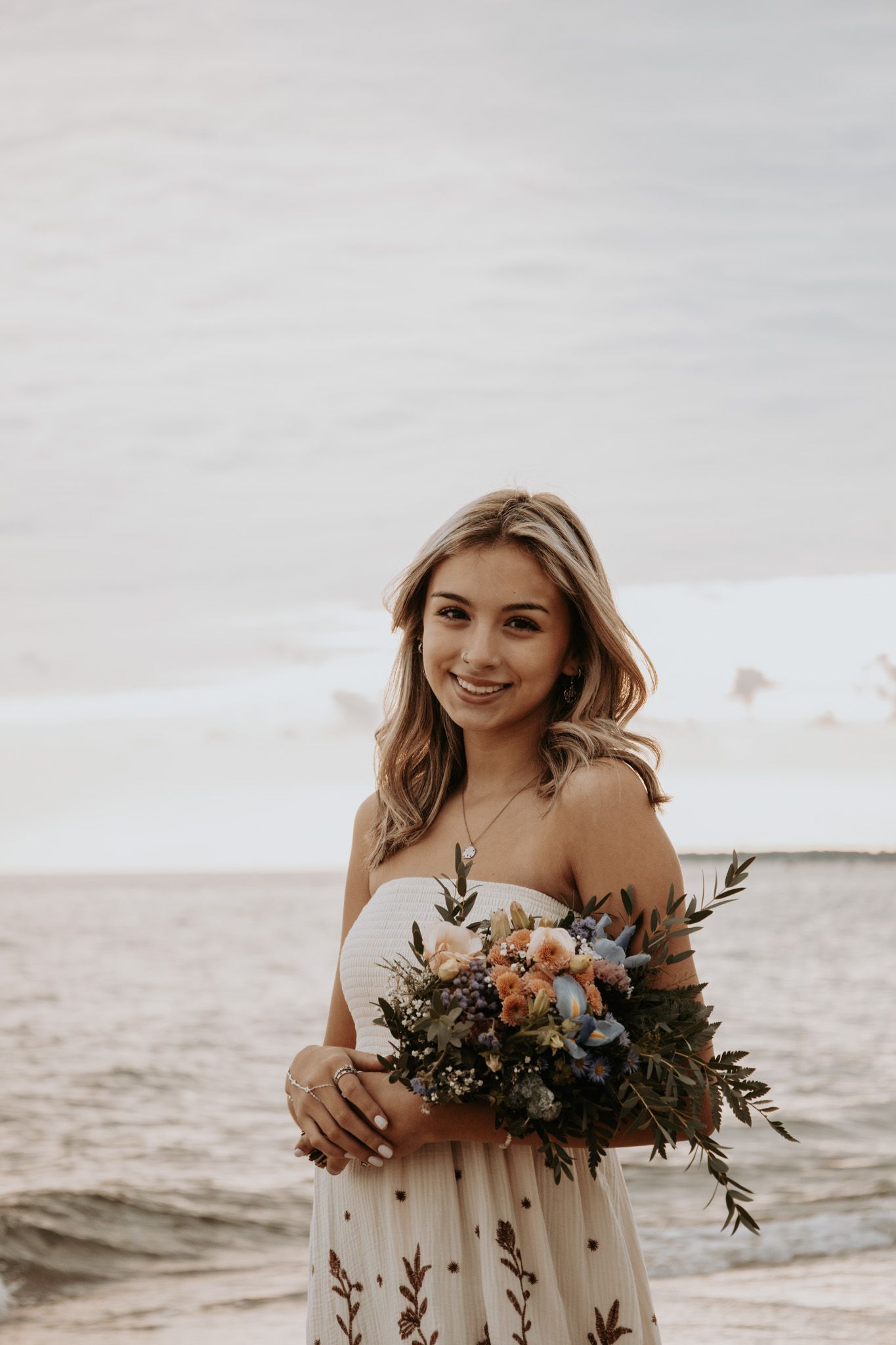 A bride in an off-shoulder white dress holds a rustic bouquet of flowers while standing on a beach at sunset.