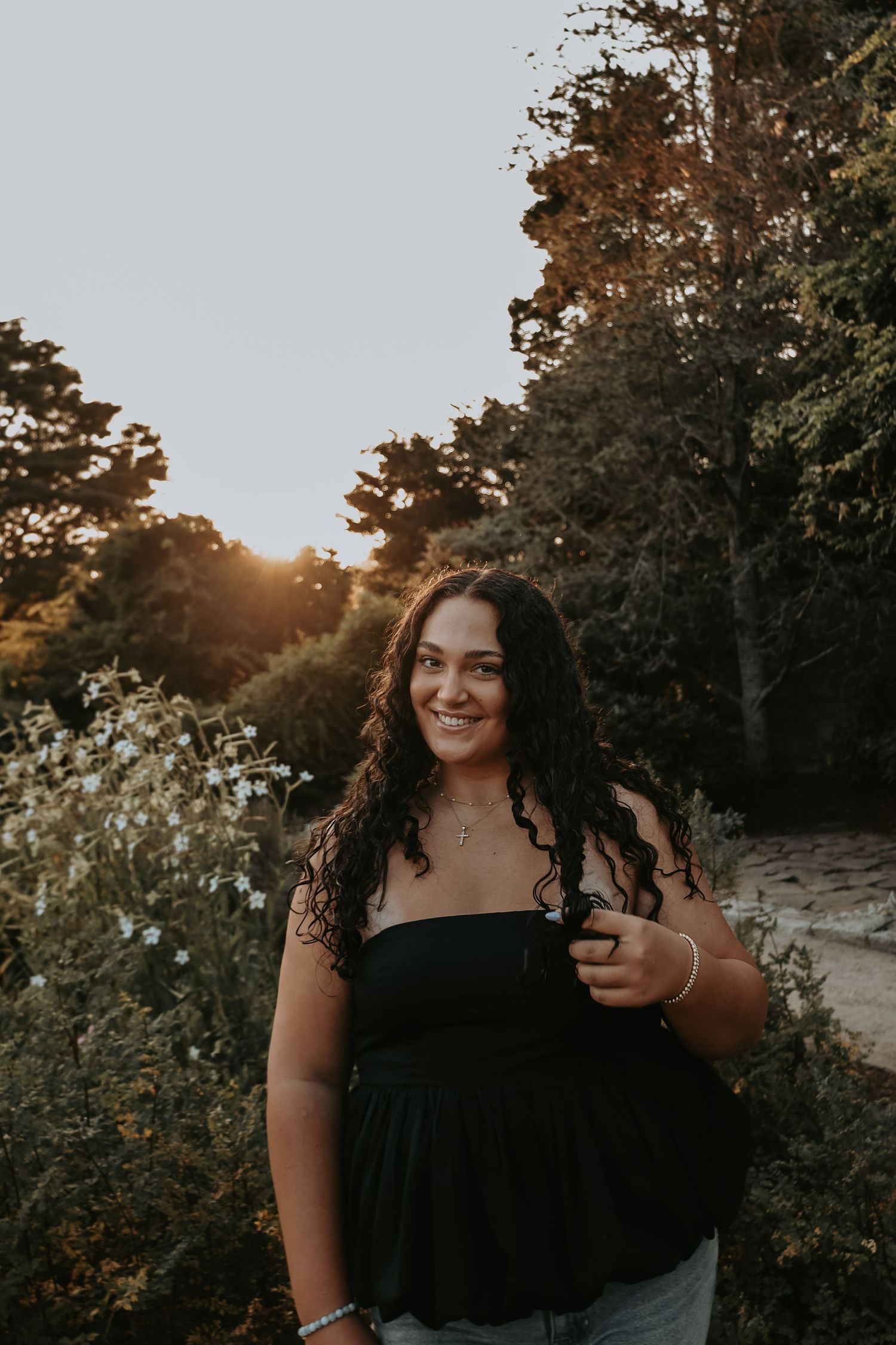A person in a black top poses outdoors against a backdrop of trees during golden hour sunset.