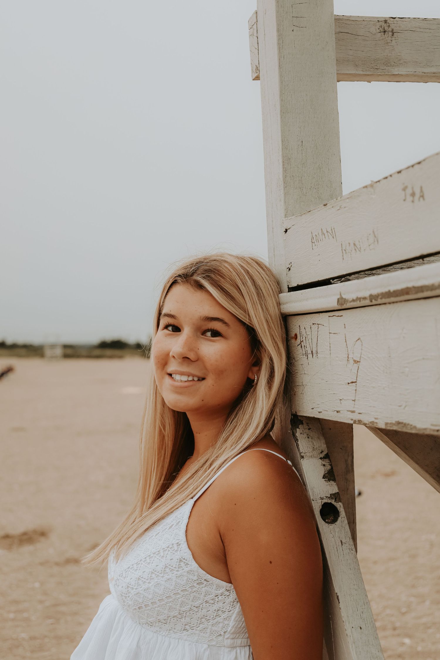 A person in a white lace dress smiles while leaning against a rustic wooden structure on a beach.