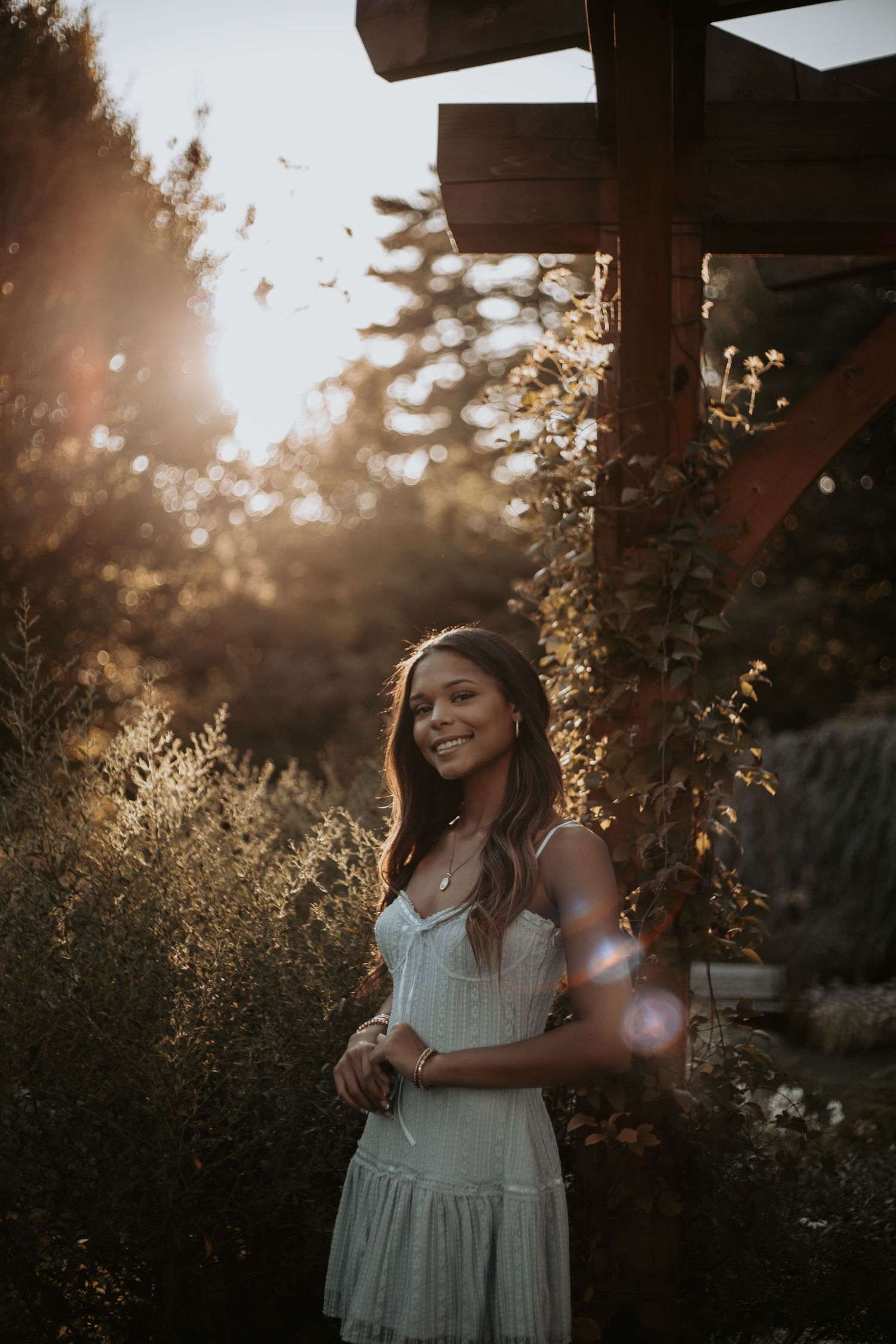 A white sundress glows in golden sunlight beneath a wooden pergola during sunset in a garden setting.