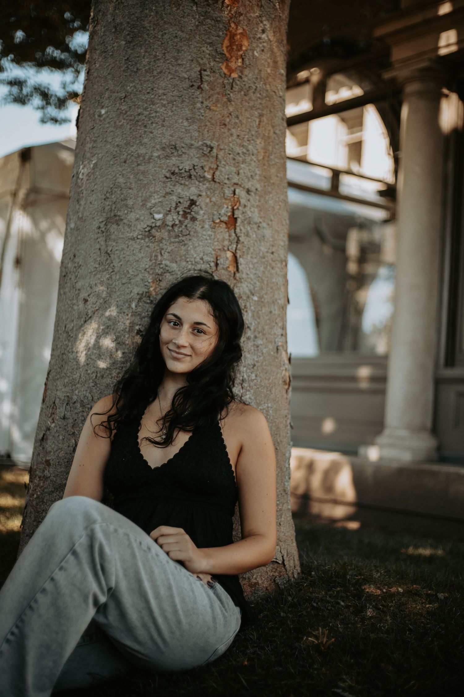 A person in a gray outfit sits against a tree trunk in warm afternoon light during an outdoor portrait session.