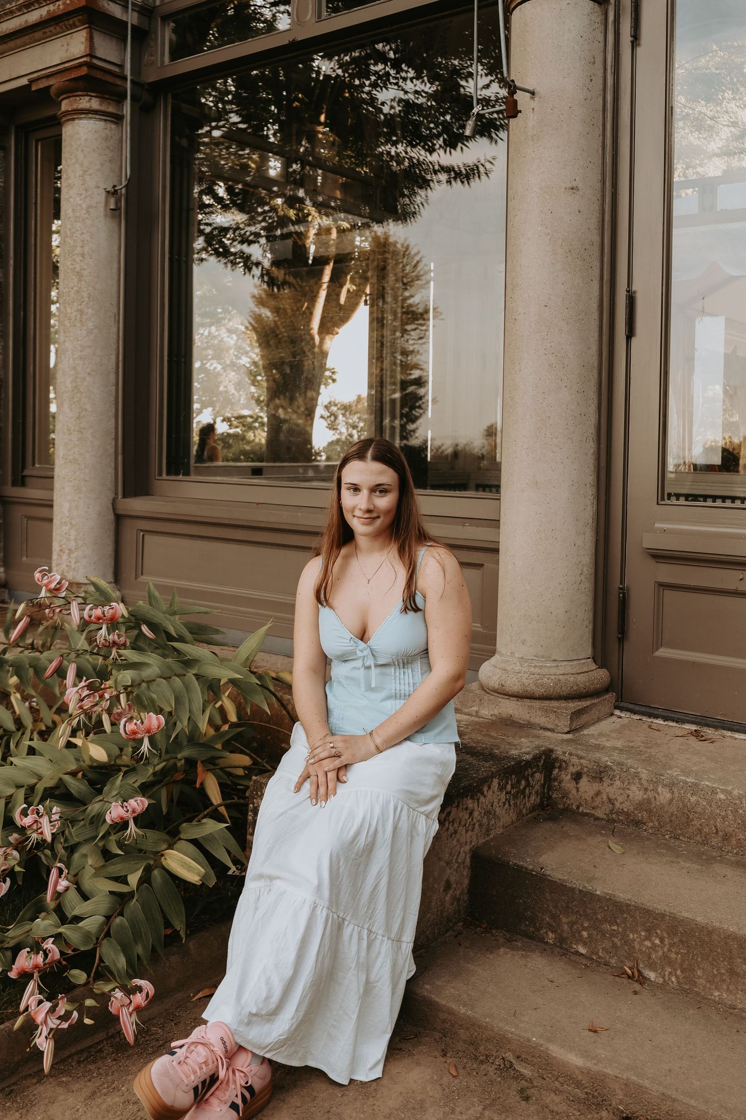 A person in a light blue dress sits on stone steps near elegant columns and pink lilies in warm evening light.