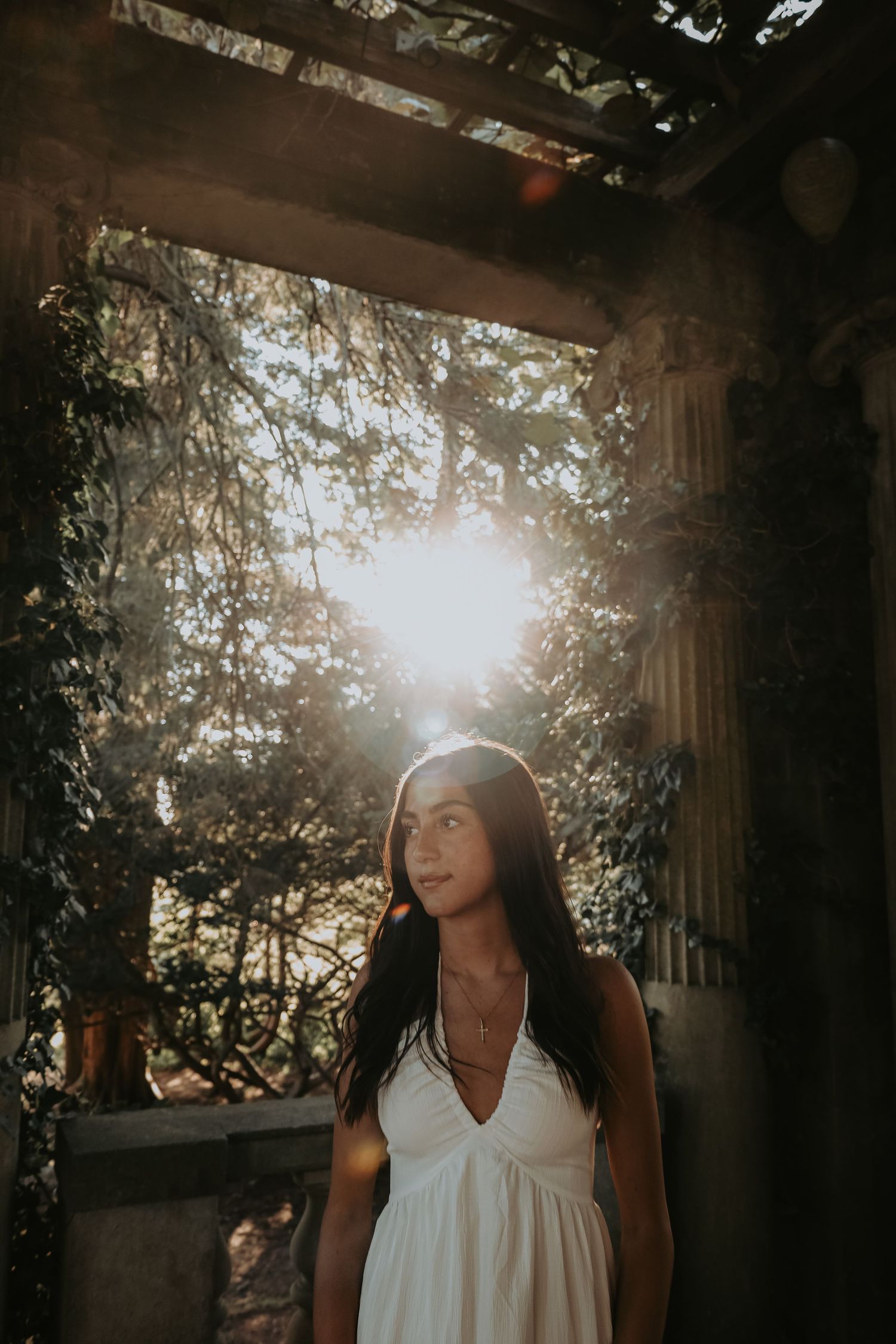 A person in a white dress stands in a sunlit garden archway creating a dreamy backlit silhouette effect.