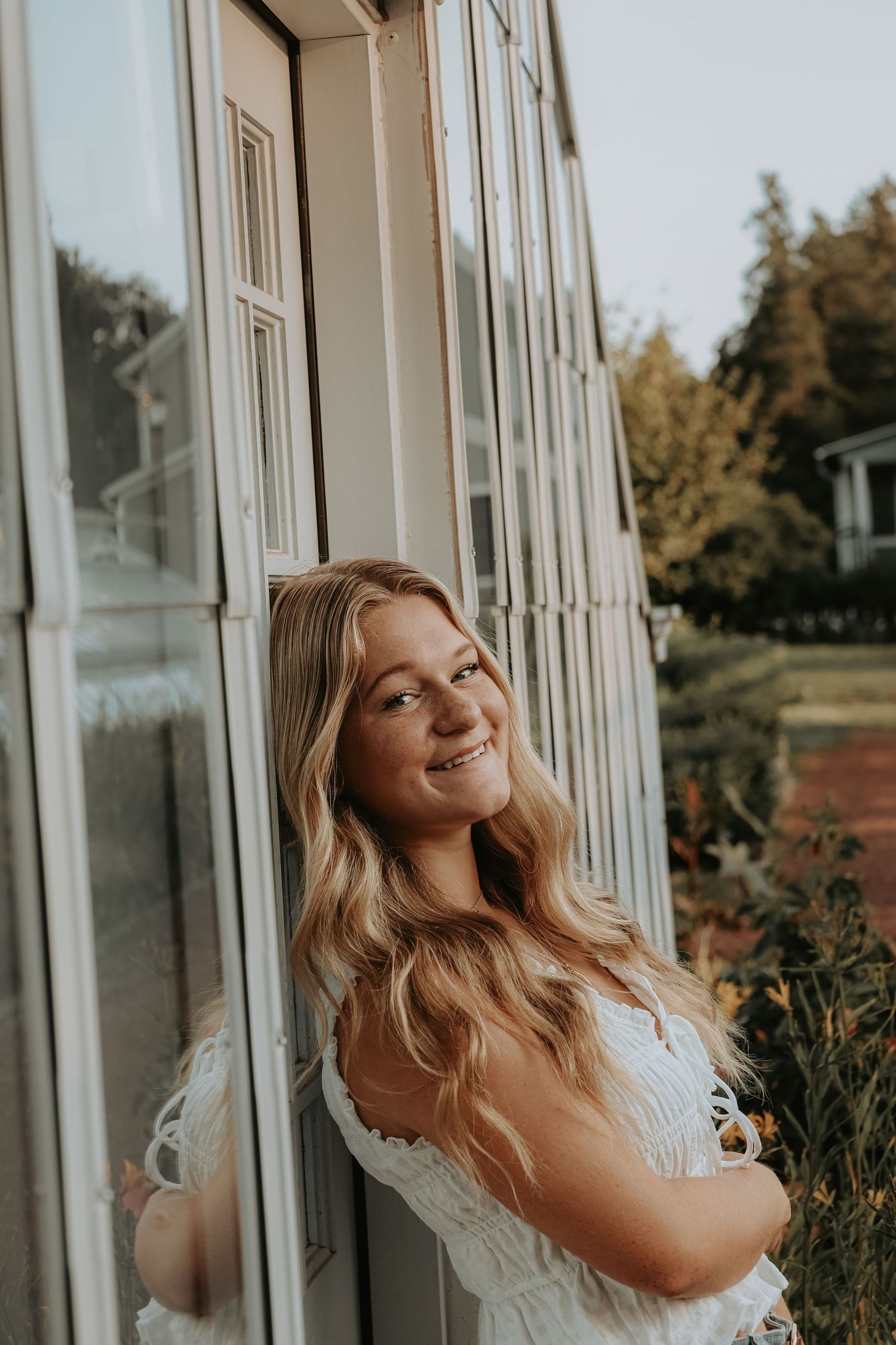 Woman in white dress poses playfully against white window frames outdoors.