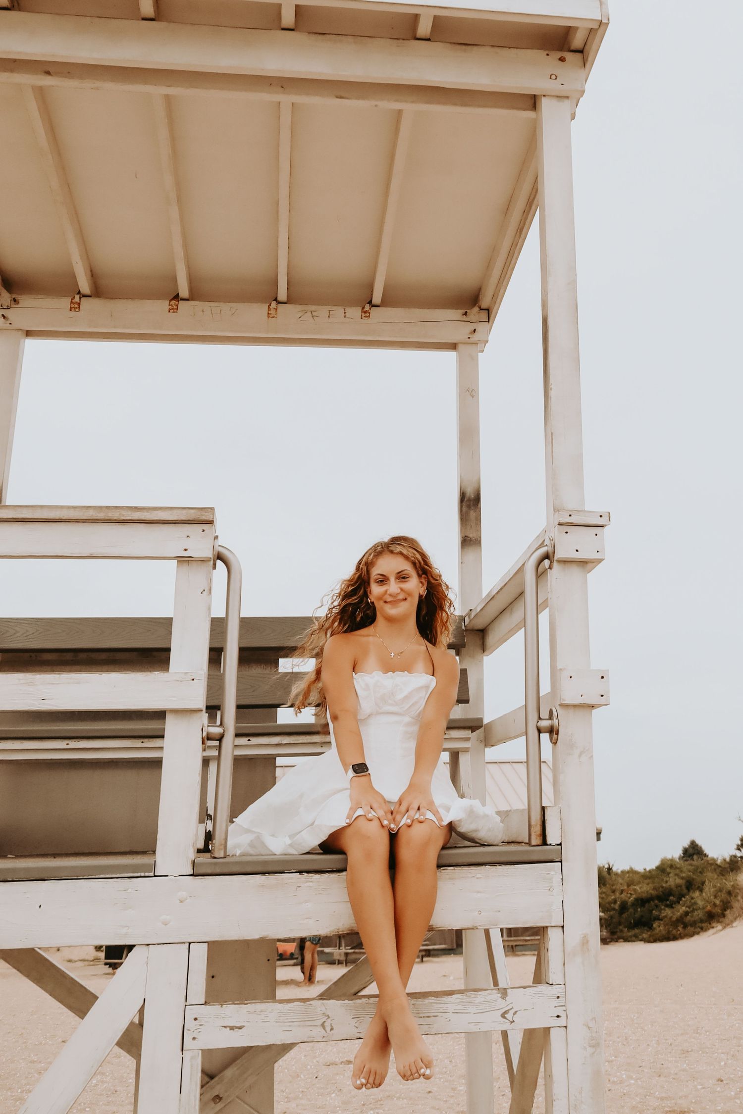 Woman in white swimsuit poses on wooden lifeguard tower at beach in warm sunlight.