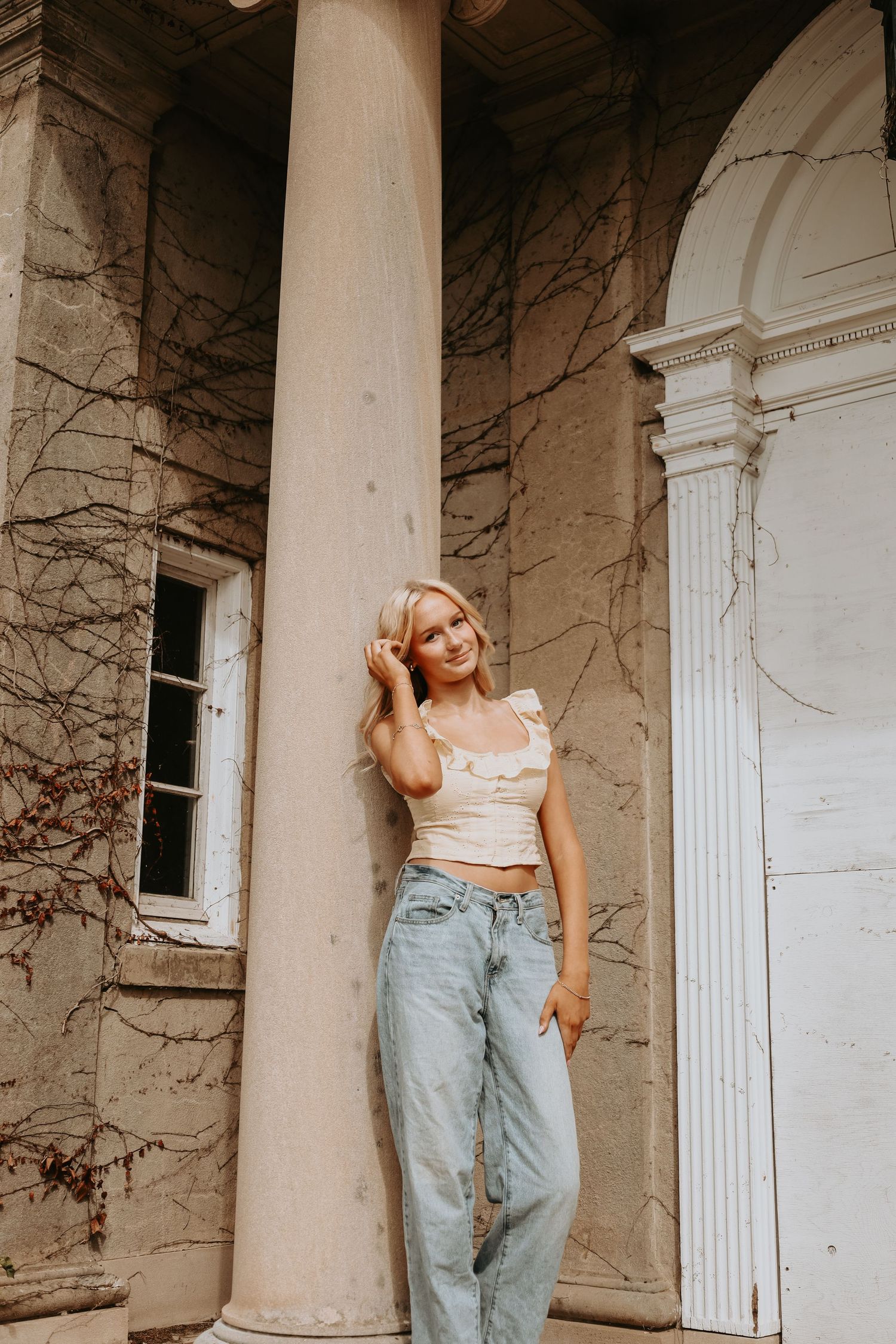 A person in light denim jeans and crop top poses casually against white columns of a building exterior.