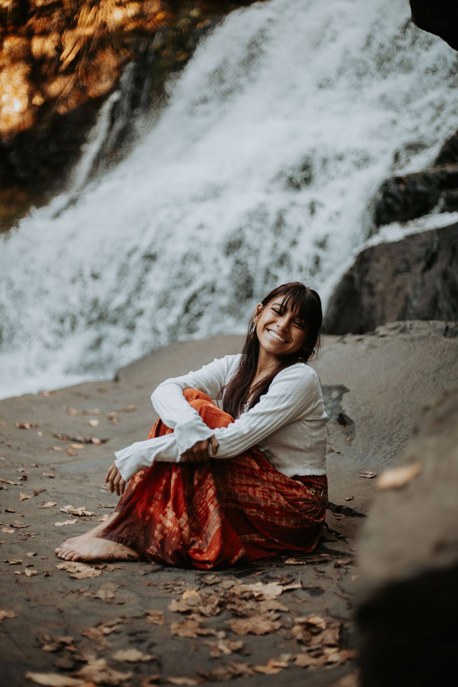 Person in white sweater and red patterned skirt poses gracefully near a waterfall with autumn leaves on the ground.