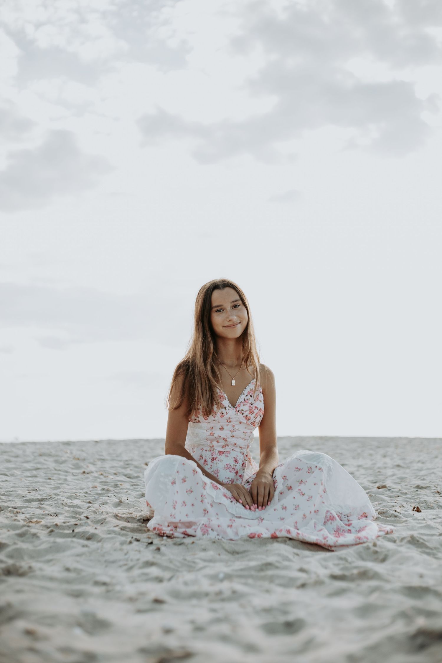 A person in a white floral dress sits gracefully on a sandy beach with the ocean visible in the background.