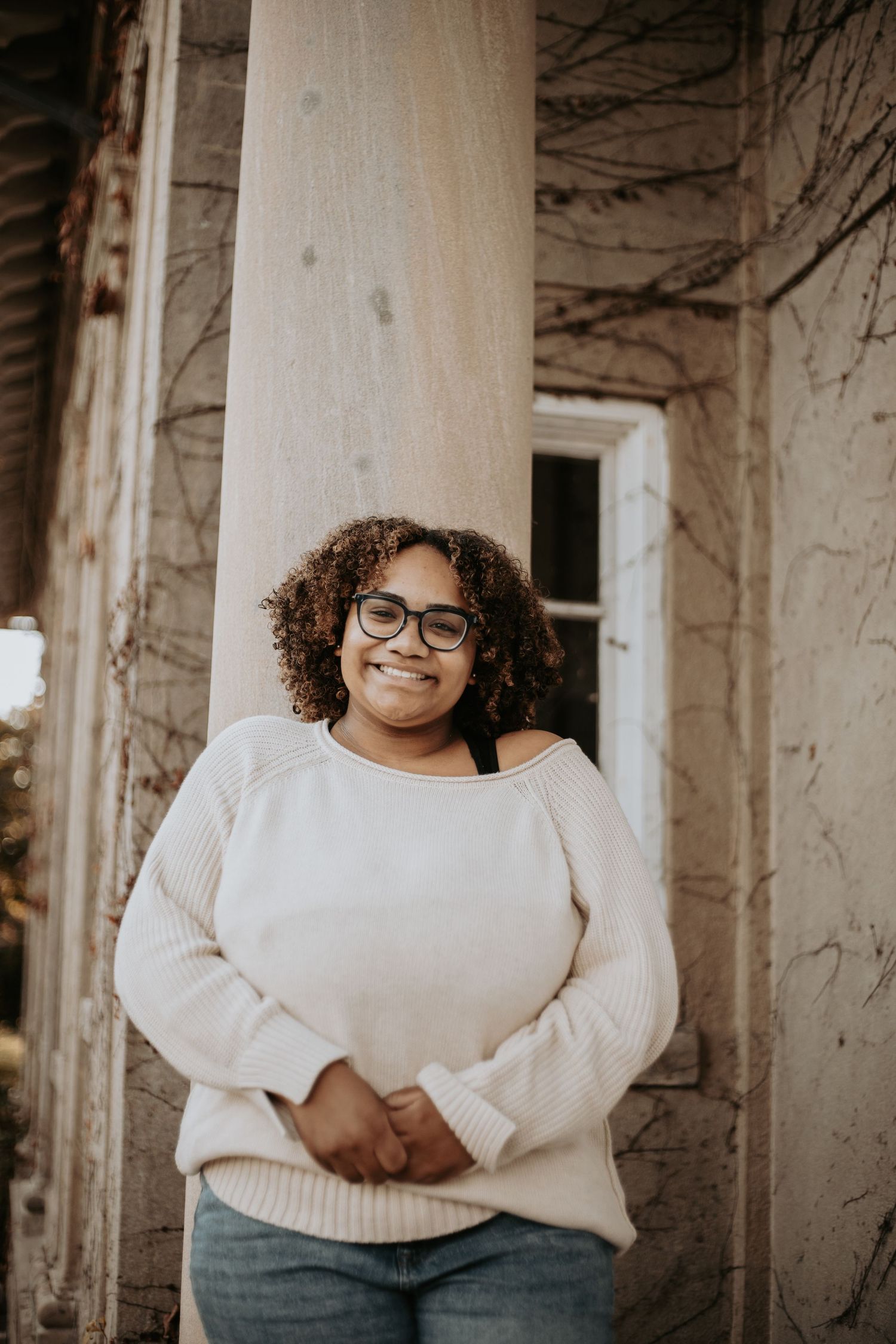 A person wearing glasses and a cream sweater smiles while standing against a rustic building exterior.