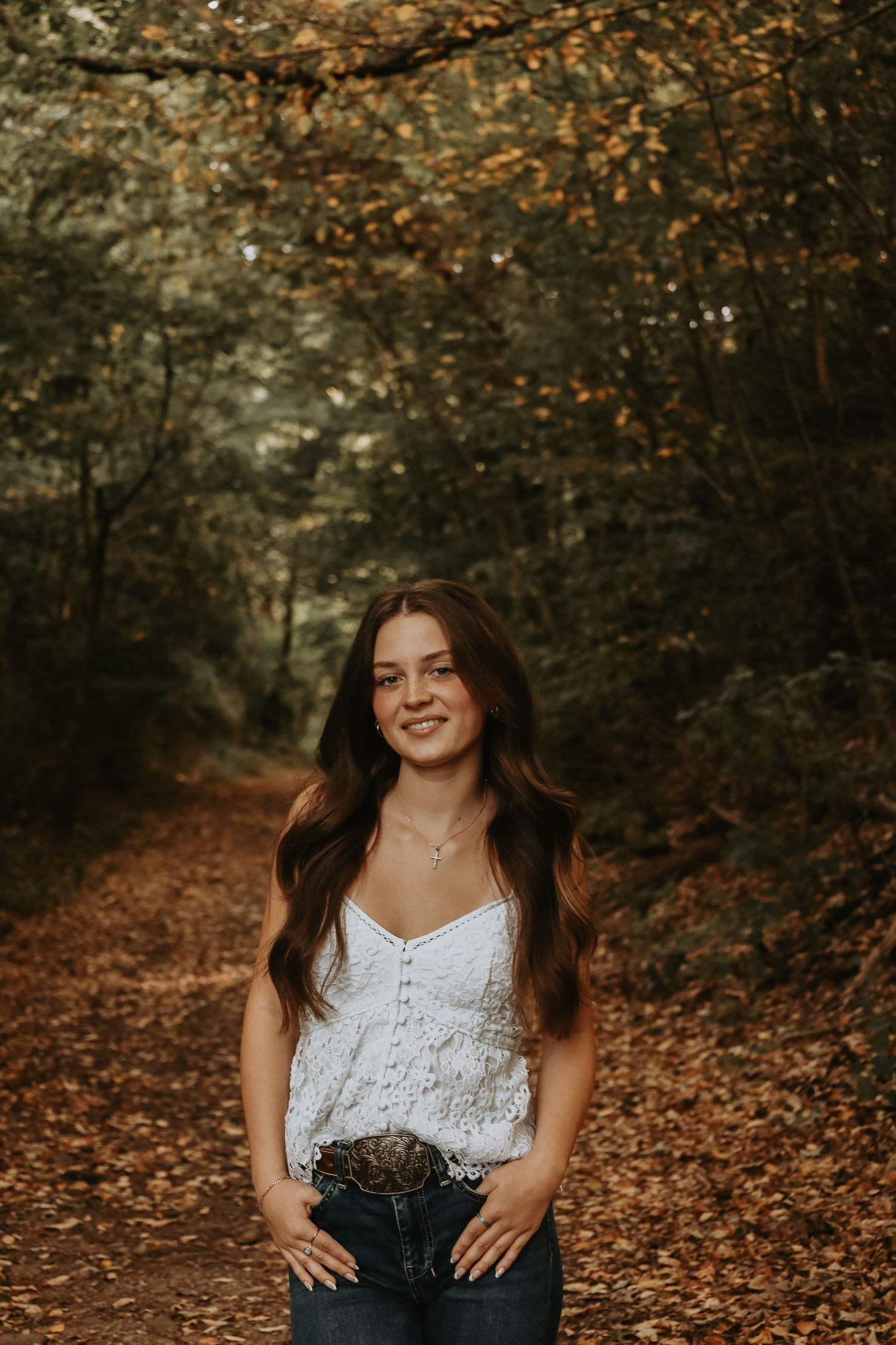 A white lace top and jeans outfit photographed on a forest path covered in autumn leaves during golden hour.