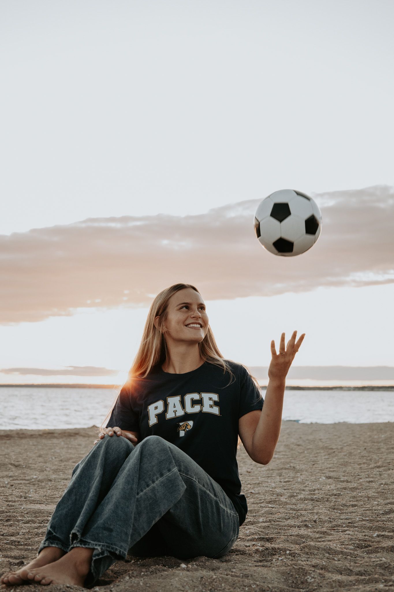 Soccer player sitting on beach tossing ball in air during sunset with ocean and mountains in background.