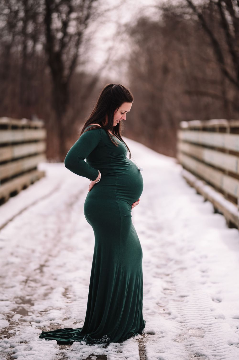 Winter maternity photo on wooden bridge featuring dark green fitted dress in snow.