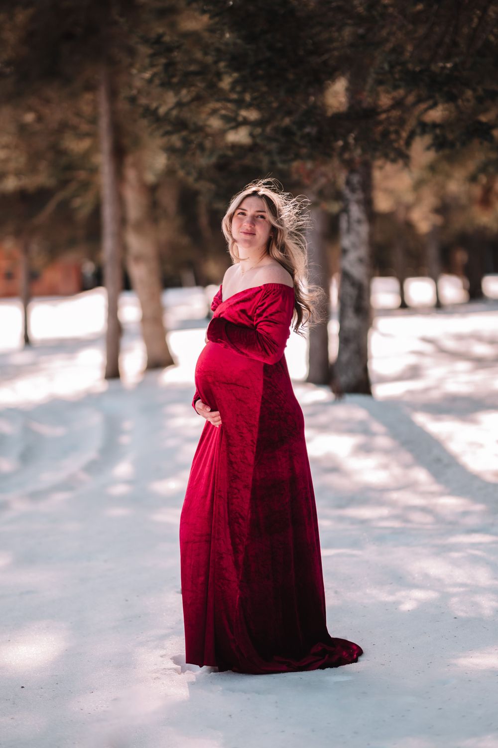 Winter maternity photo featuring red velvet dress in snowy forest setting.