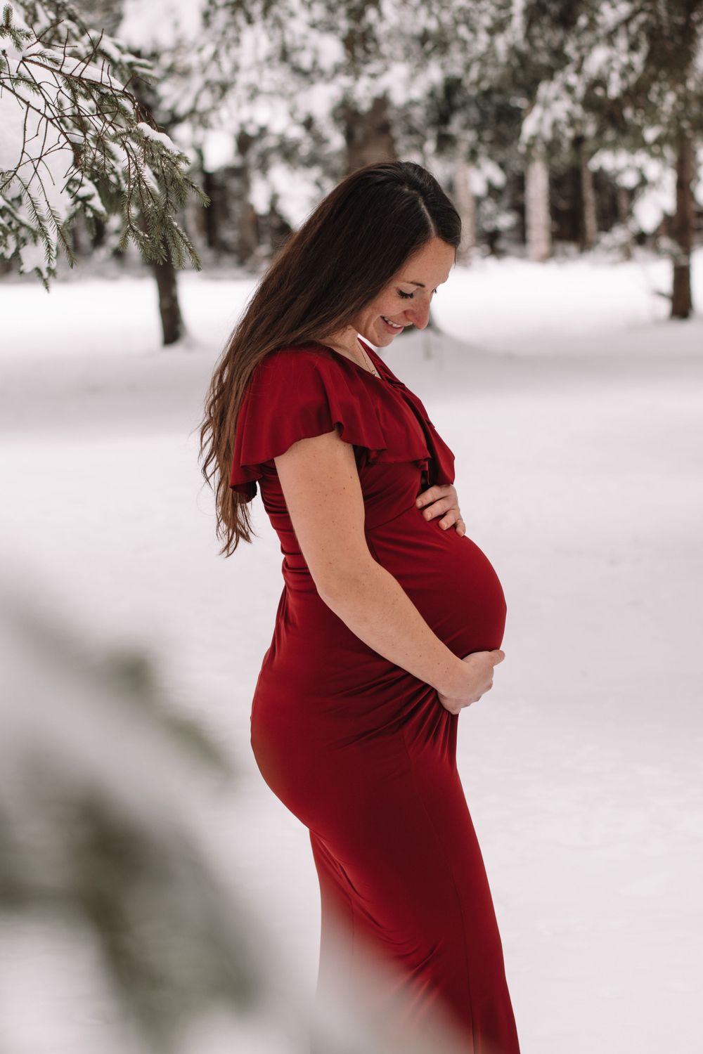 Maternity portrait in burgundy dress against snowy winter landscape.