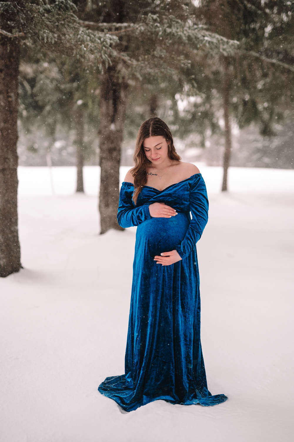 Maternity portrait of woman in blue velvet dress standing in snowy forest setting.