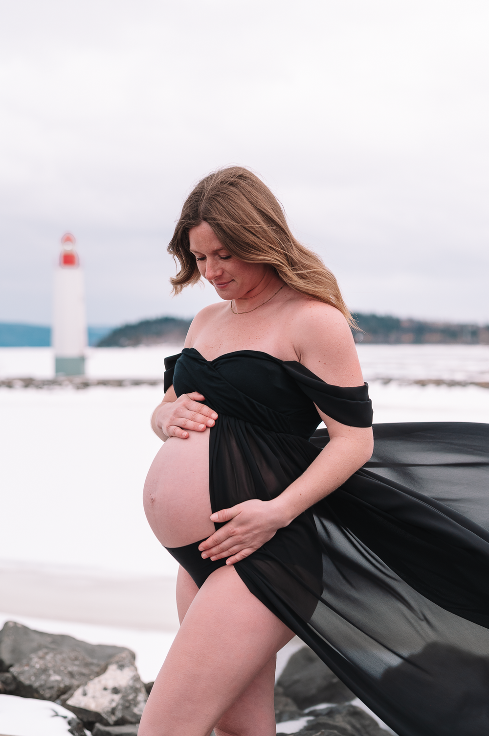 A maternity photo shoot at a lighthouse features an expectant mother in a flowing black dress against a coastal backdrop.