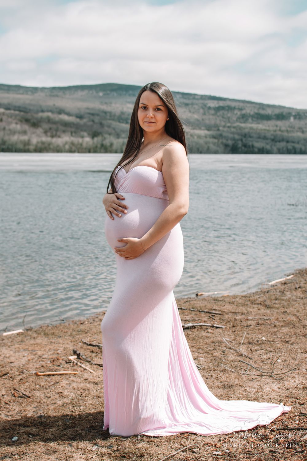 Pregnant woman in pink dress poses by lakeside during maternity photoshoot.