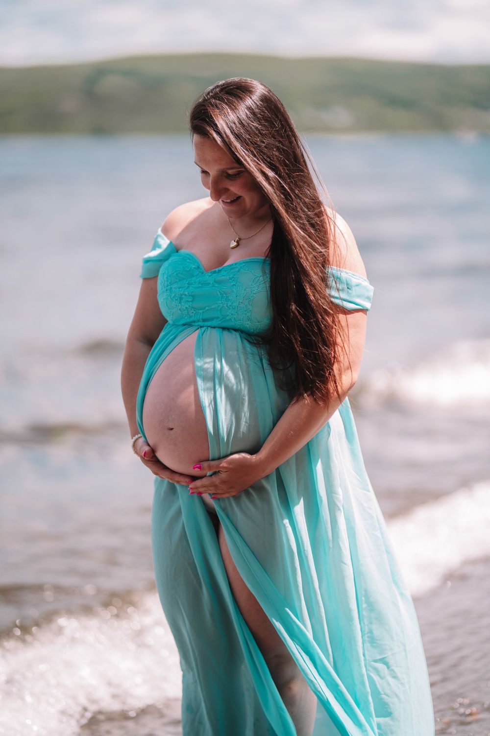Beachside maternity photo with flowing turquoise dress and waves in background.
