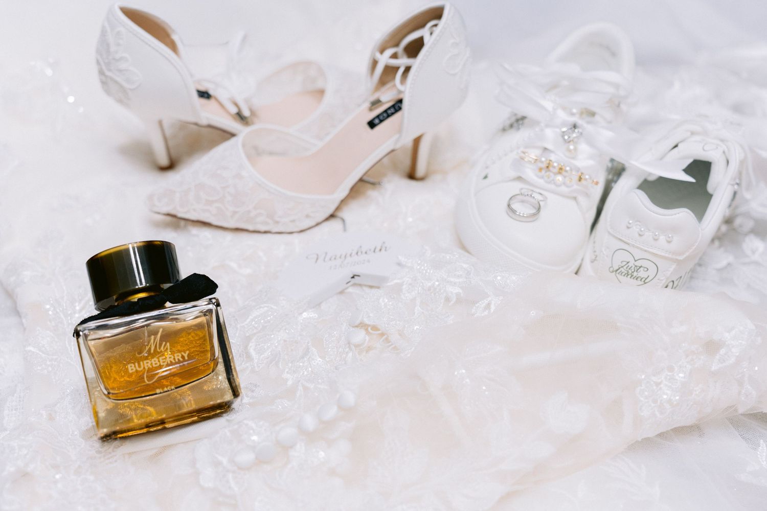 Flat-lay of bridal wedding details, including lace heels, custom sneakers with pearl accents, a perfume bottle and wedding rings.