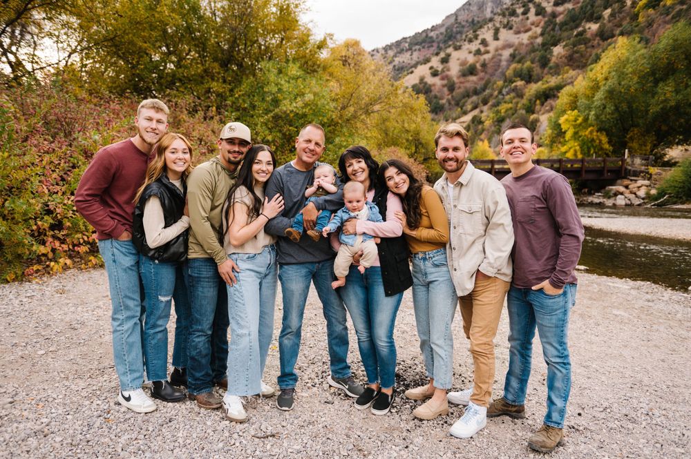 Extended family group smiling together on gravel path with mountains and fall foliage behind.