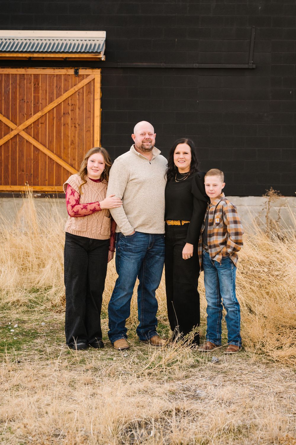 A family of four poses for a portrait in front of a rustic barn with wooden details.