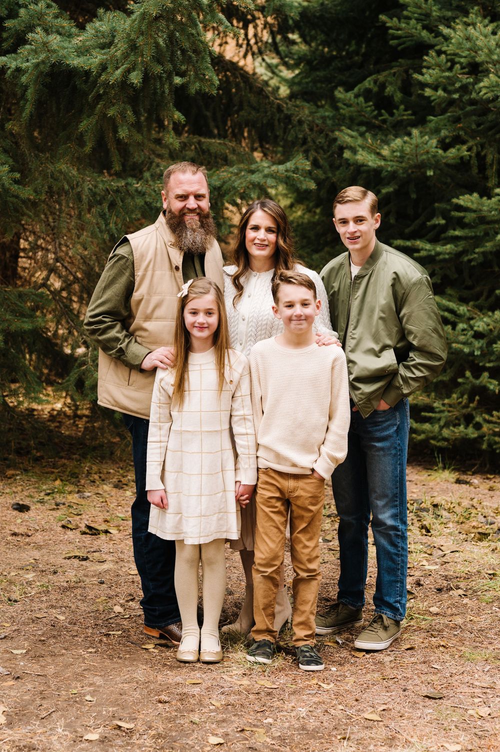 Family of four posing together in front of evergreen trees in casual attire.