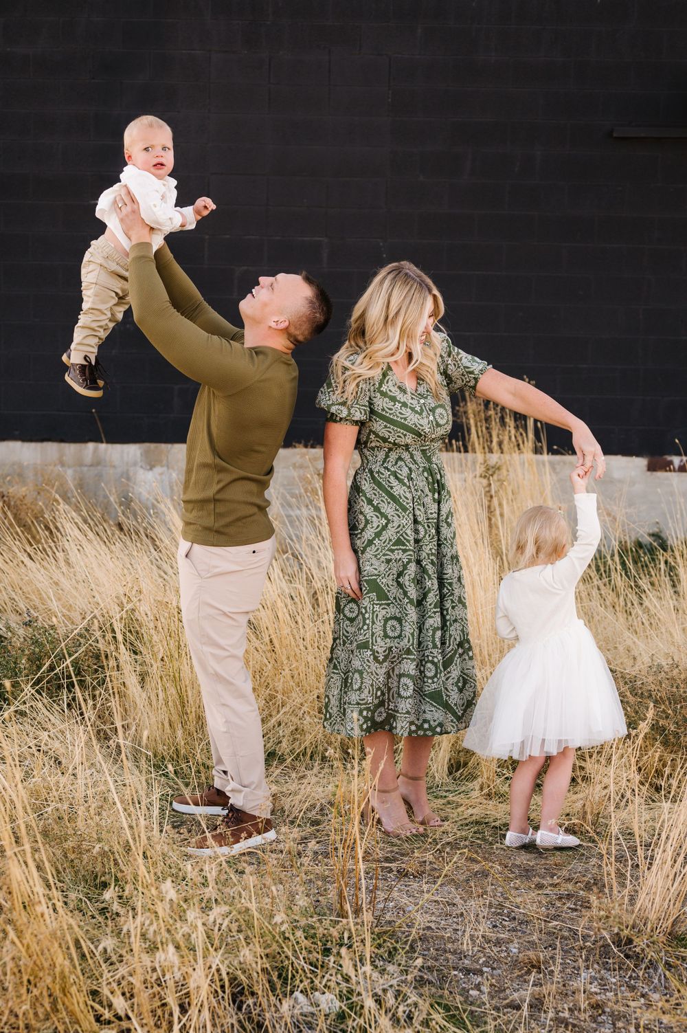 A family plays together in a field, with parents lifting children against a dark wall backdrop.