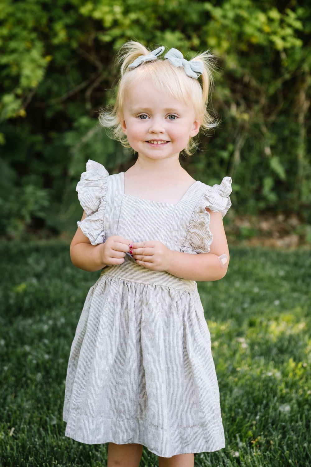 A young child in a white ruffled dress standing on grass with natural greenery in background.