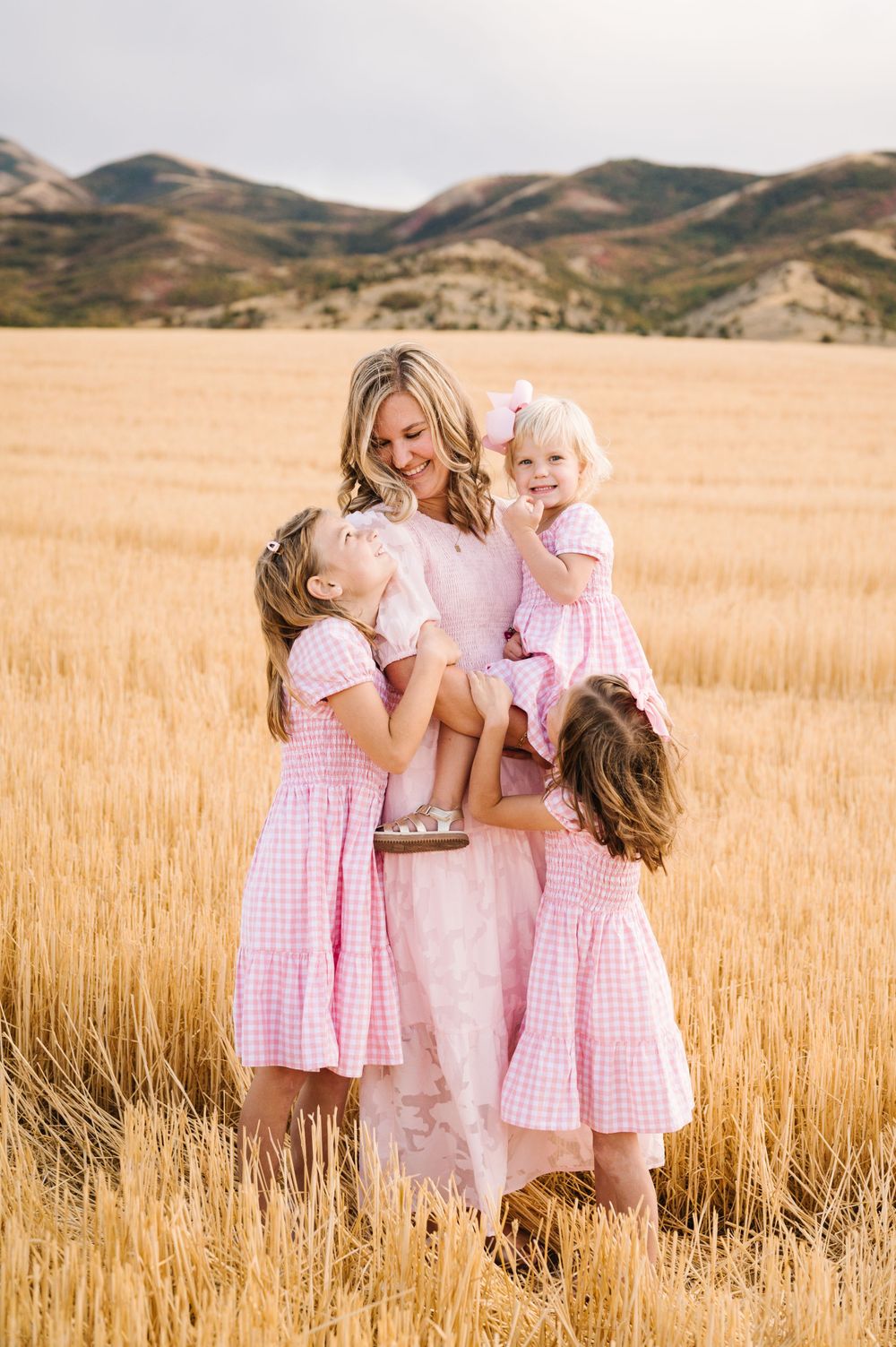 Group wearing pink gingham dresses poses together in golden field with mountains behind.