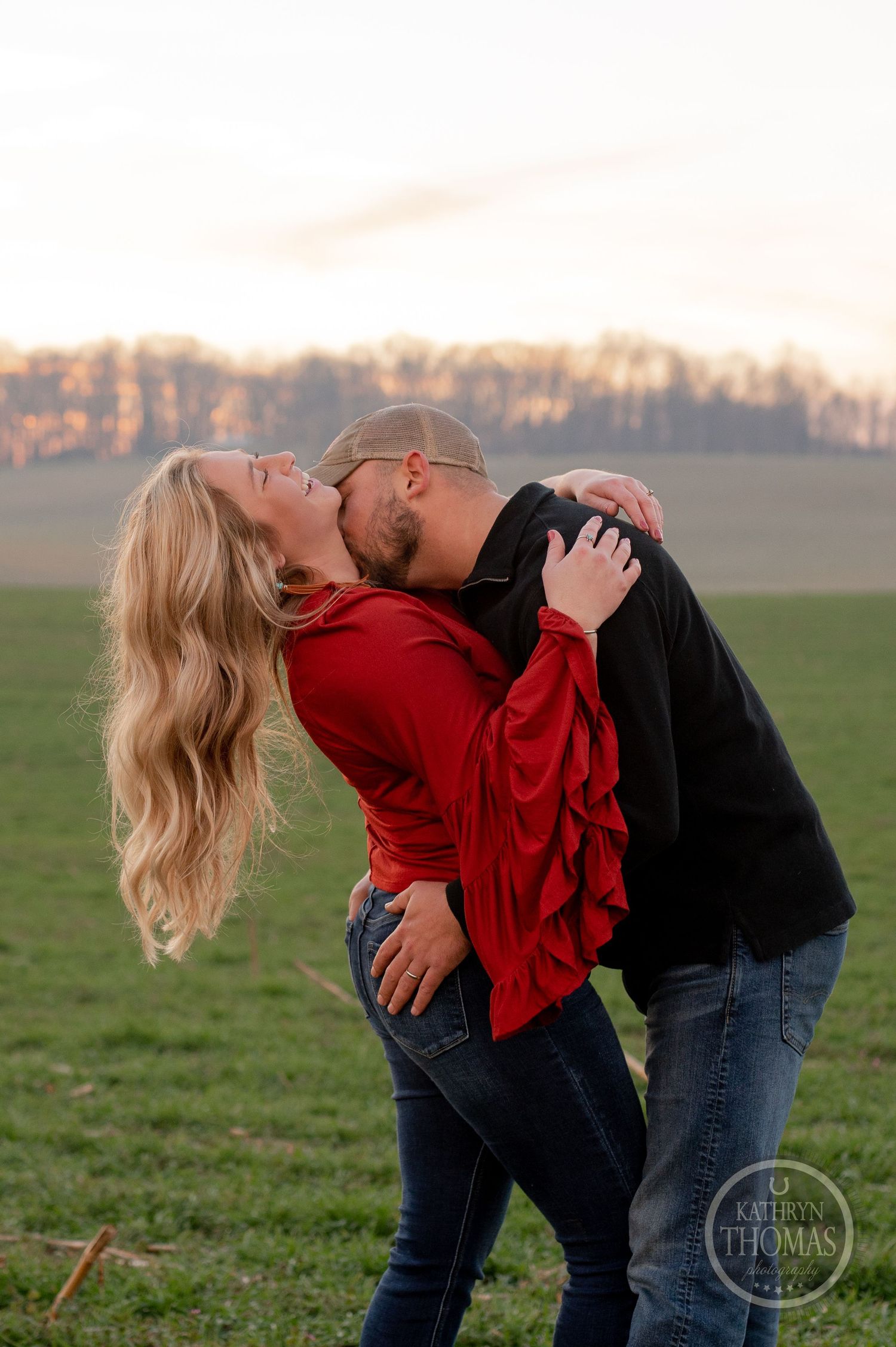 A couple embraces in a romantic pose against a rural sunset backdrop.