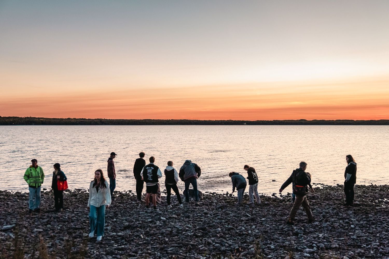 Silhouettes of people standing on rocky beach at sunset with orange sky reflecting on water.