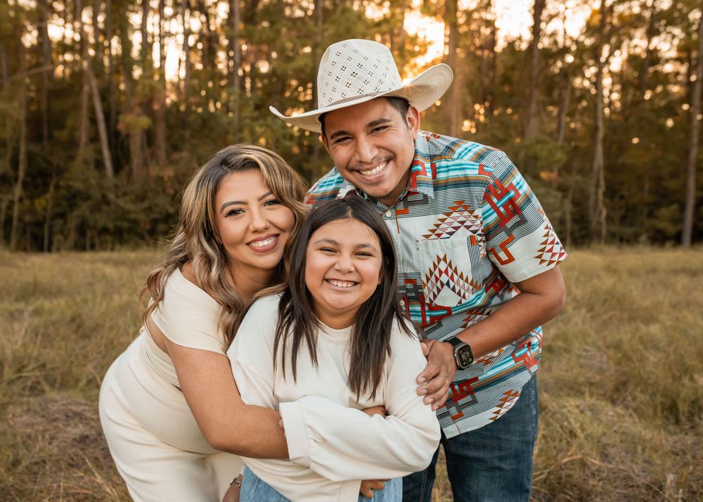 Three people in casual attire embrace and smile while posing together in a grassy field at sunset with pine trees behind them.