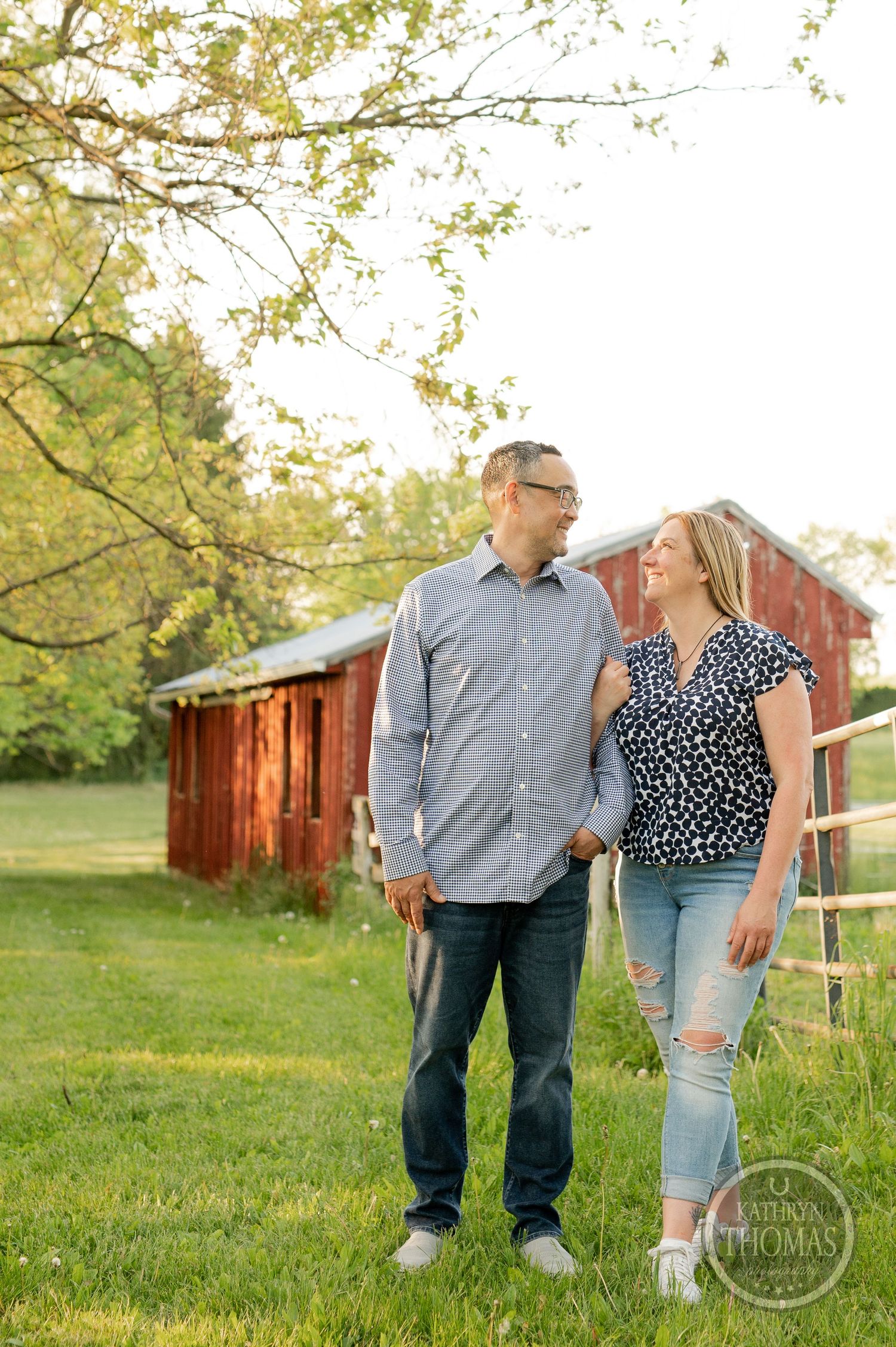 A couple enjoys a tender moment outdoors near a rustic red barn and wooden fence in springtime.