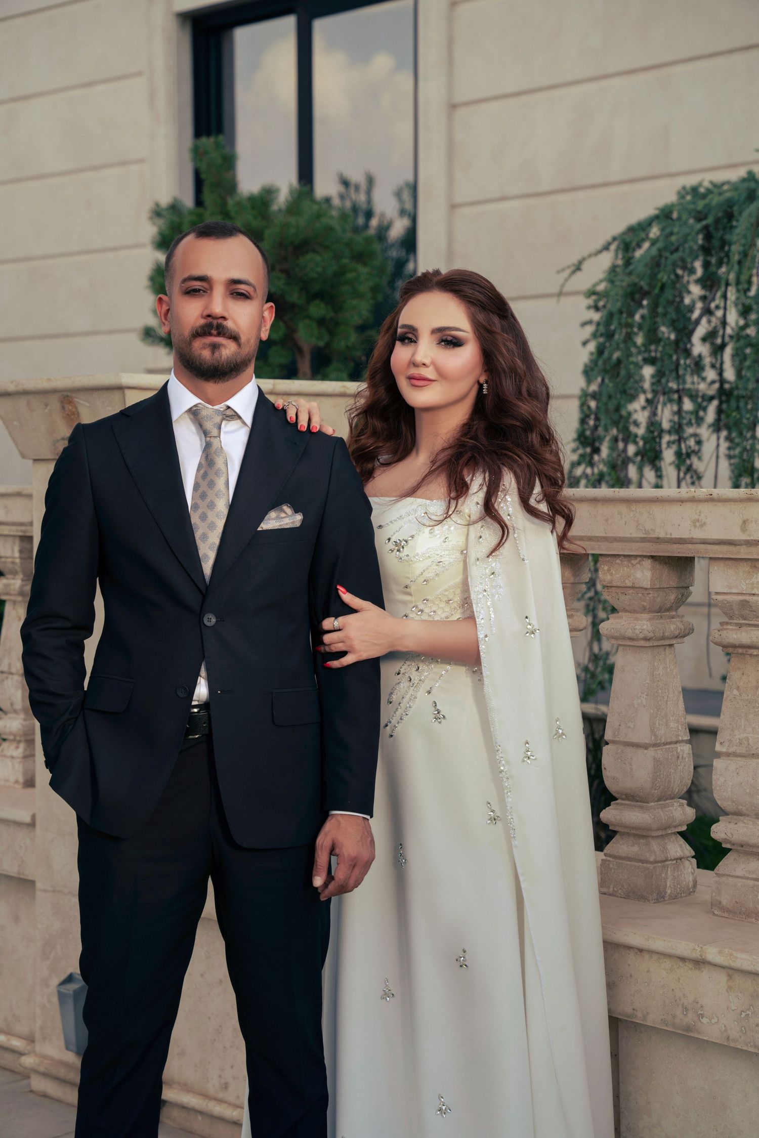 A couple poses together in elegant wedding attire of a black suit and flowing white gown with embellishments.