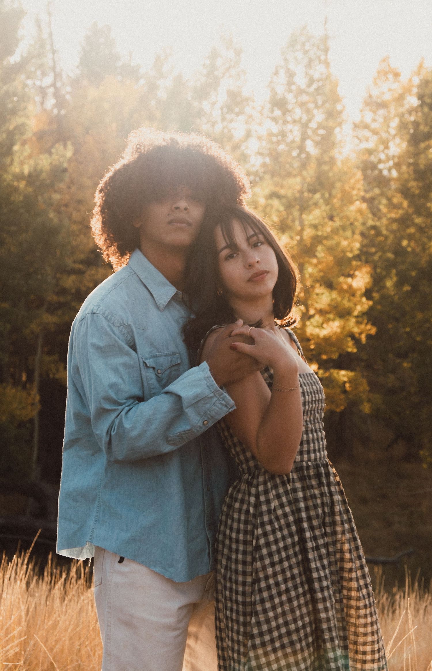 A couple embraces in a golden field at sunset wearing blue and plaid outfits.