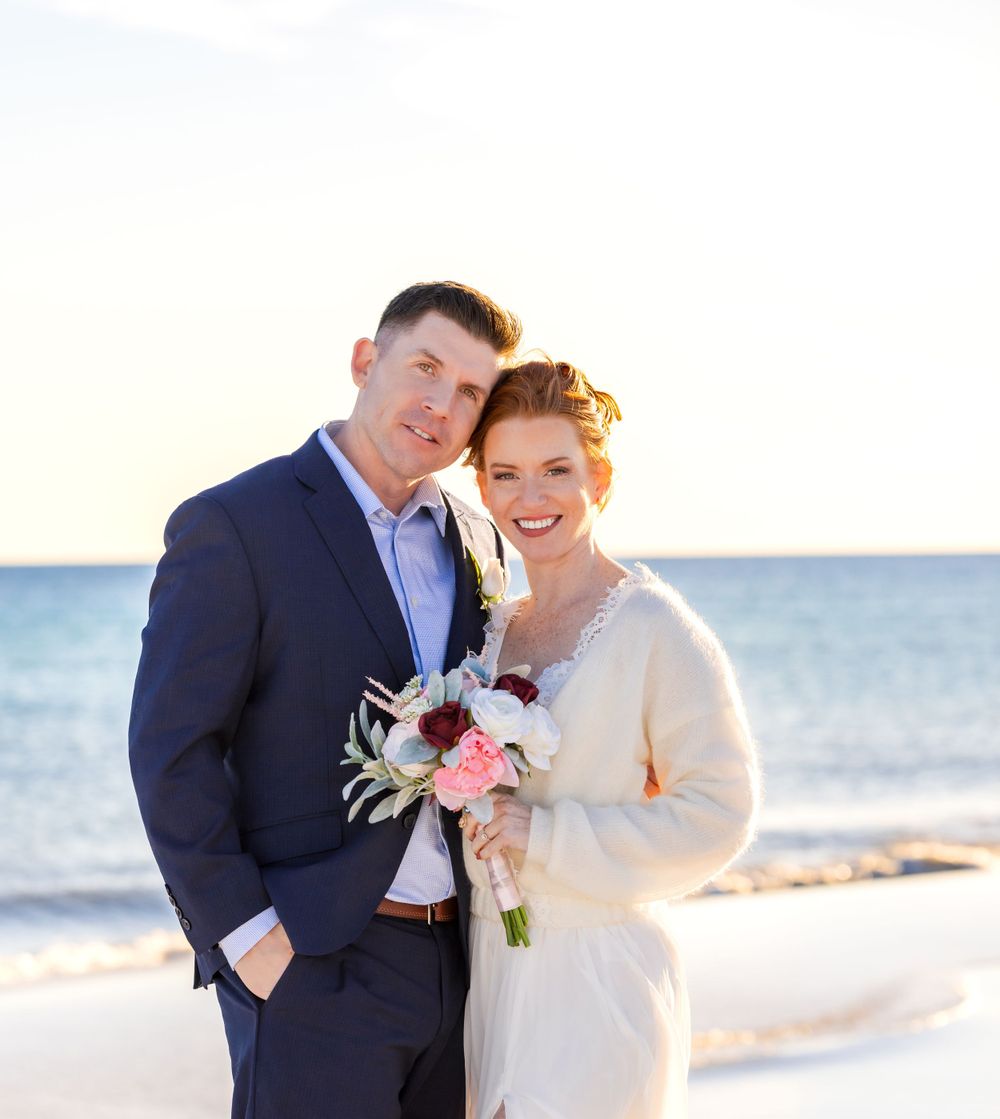 A couple embraces on a sunny beach at sunset wearing a navy suit and white dress with matching bouquet.