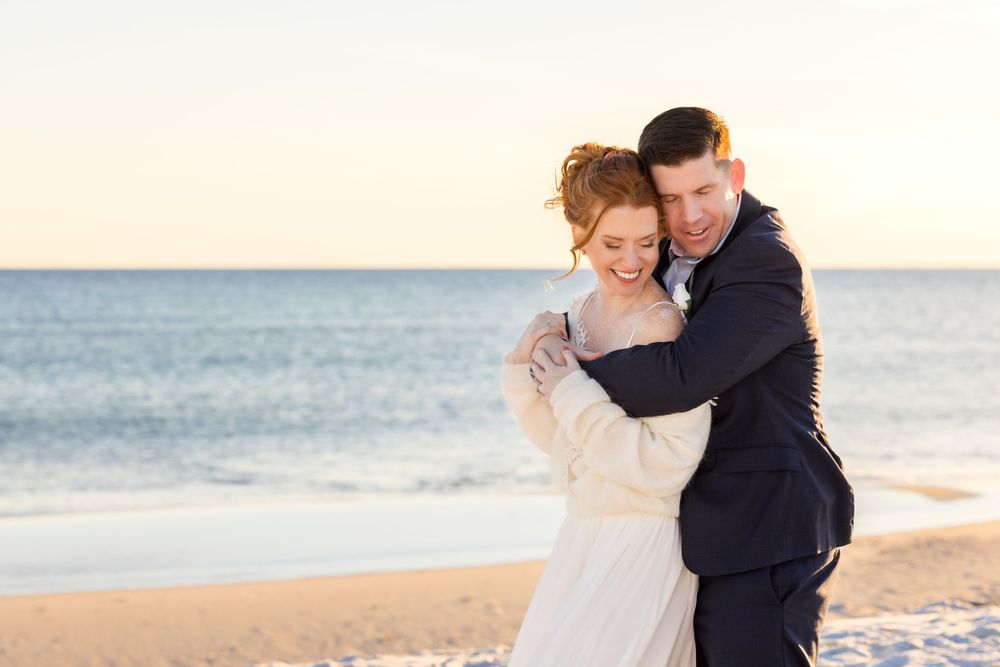 A romantic couple embraces on a sandy beach at sunset with the ocean in the background.