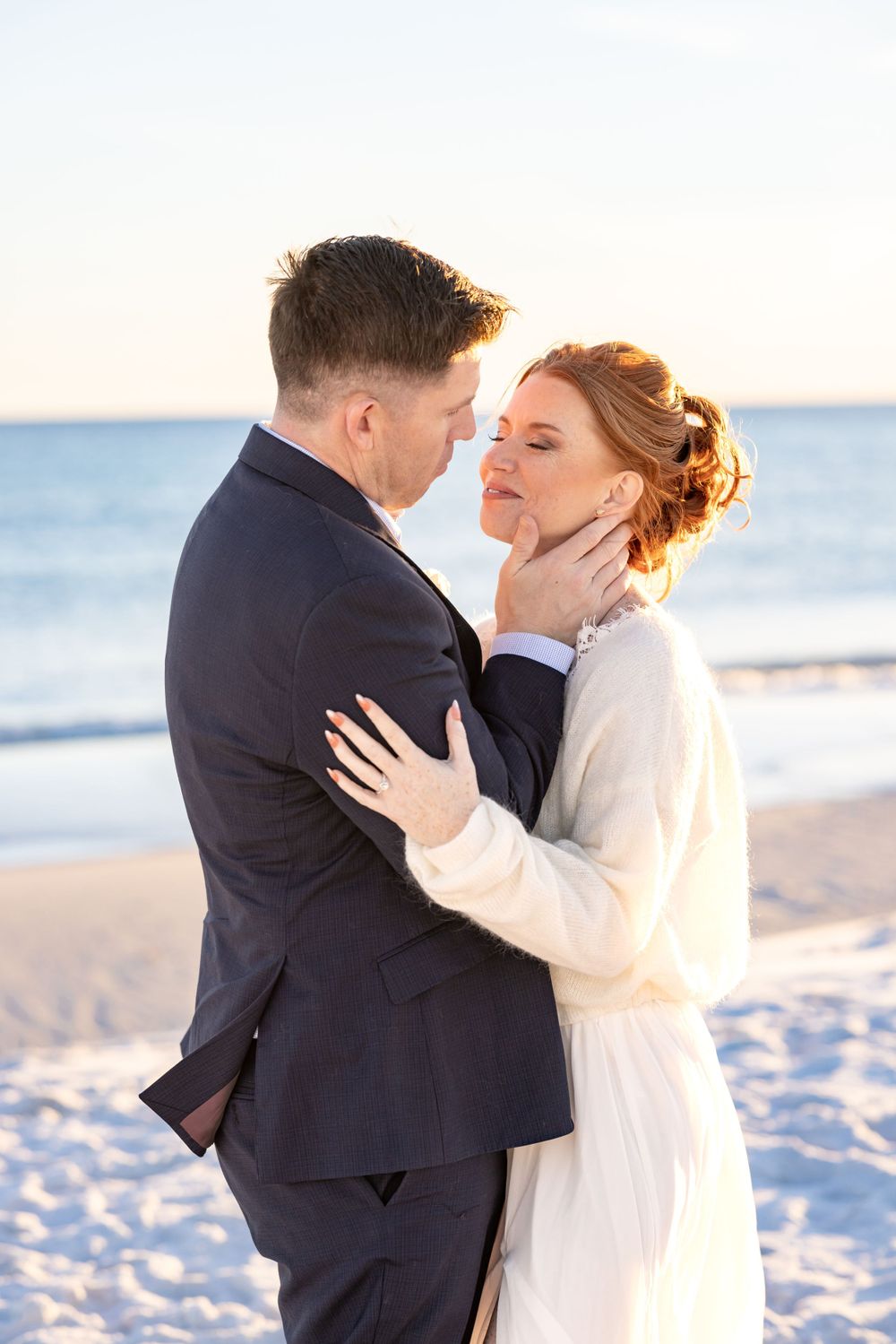 A romantic couple shares an intimate moment on a sunny beach, with the ocean and sunset in the background.