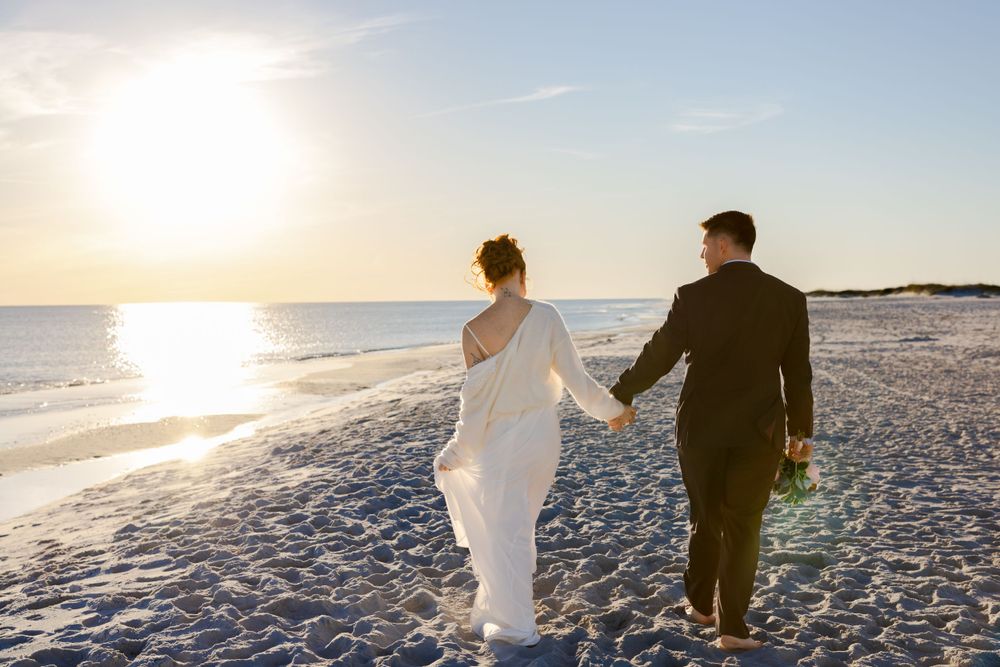 Two people walking hand in hand on a sandy beach at sunset with ocean waves and glowing sunlight in the background.