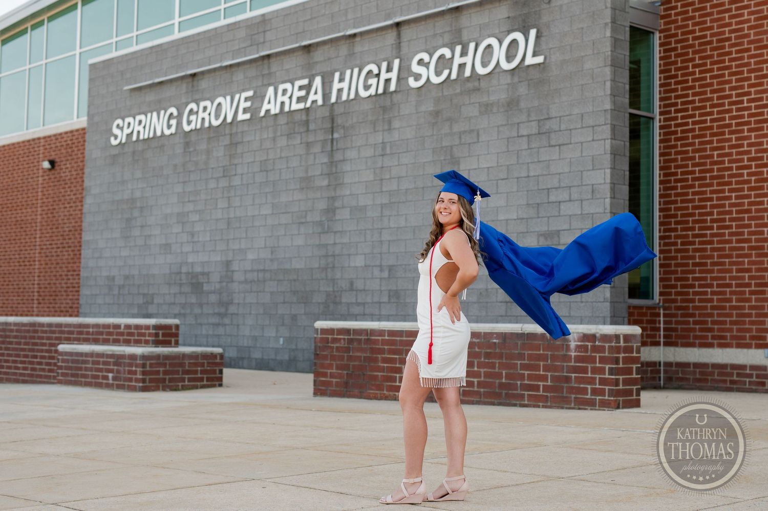 Playful graduation portrait with flowing blue gown against brick school building exterior.