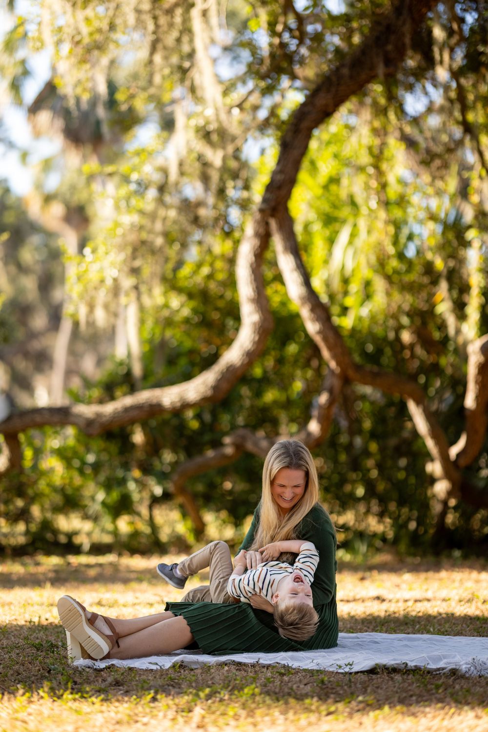 Two people relax on a blanket under trees with dappled sunlight and fallen autumn leaves in a peaceful park setting.