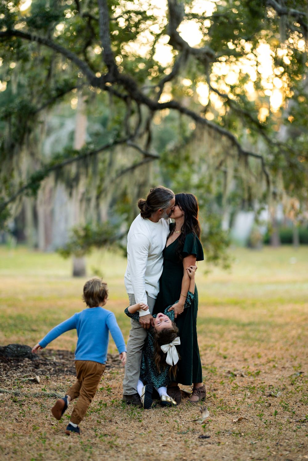 Family shares tender moment together under moss-draped trees in a sunlit park as child runs playfully nearby.