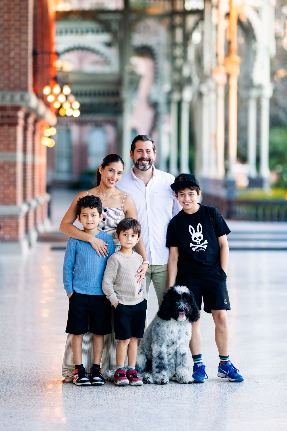 Family group stands together in a covered outdoor walkway with brick columns and glowing lights in background.