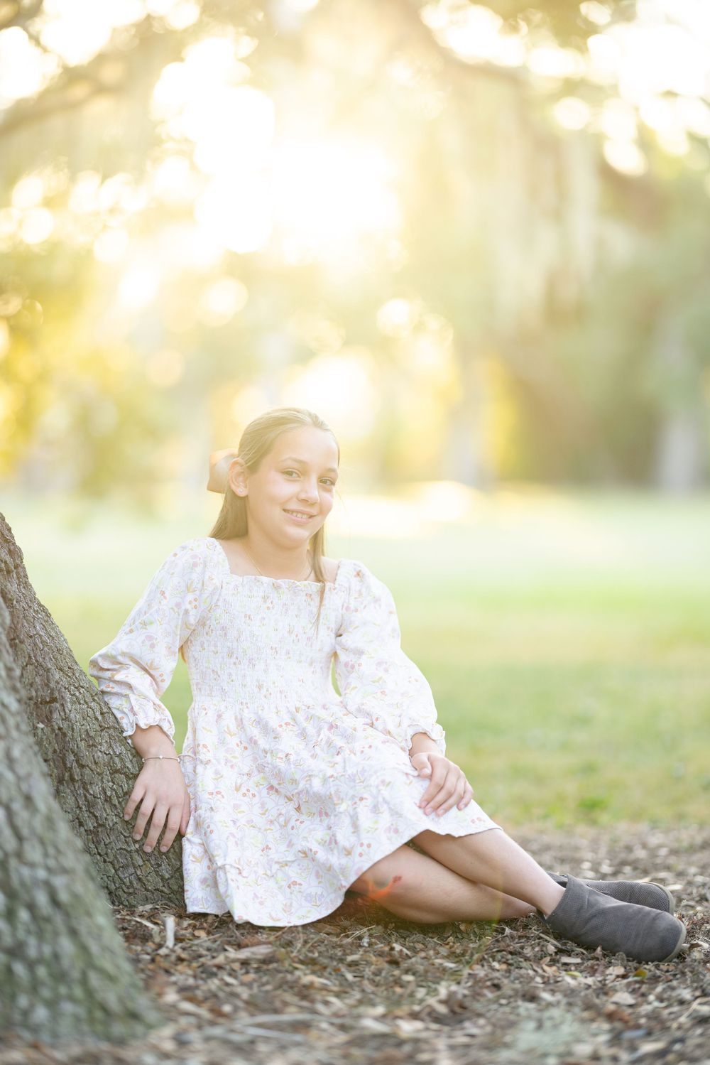 A person in a white lace dress sits on fallen leaves near a tree trunk during golden hour lighting.