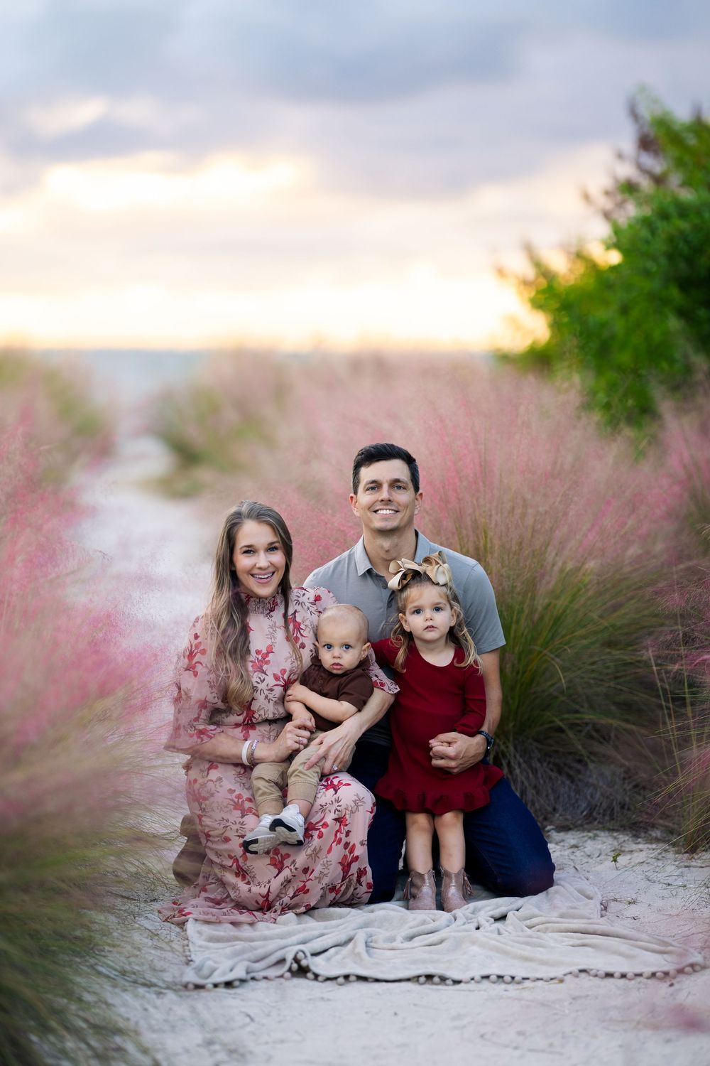 A family sits together on a sandy beach surrounded by pink muhly grass during sunset.